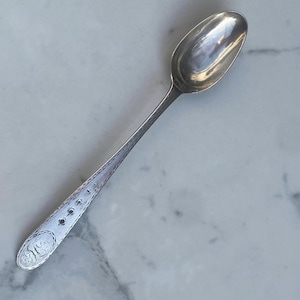 Puede incluir: Una cucharita de plata con un diseño ornamentado en el mango. La cuchara tiene un cuenco redondeado y un mango largo y delgado.