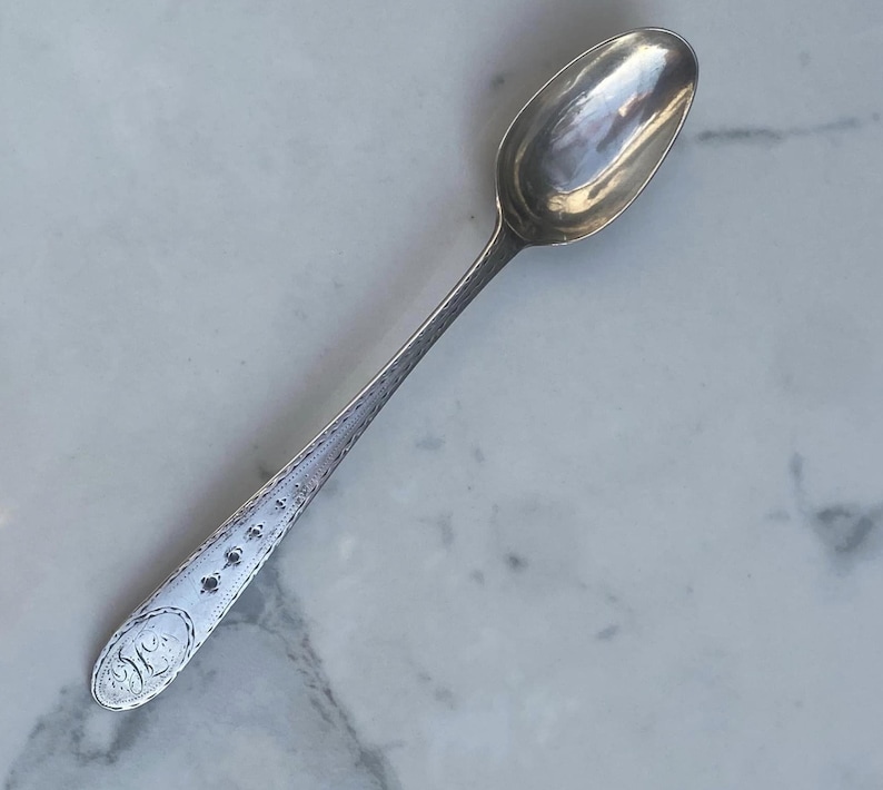 May include: A silver teaspoon with an ornate design on the handle. The spoon has a rounded bowl and a long, slender handle.