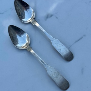 Puede incluir: Dos cucharas antiguas de plata con cuencos ovalados y mangos largos y delgados. Cada mango está grabado con una inicial en escritura. Las cucharas se muestran sobre una superficie clara.