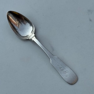 May include: A silver spoon with a rounded bowl and a long, flat handle. The handle is engraved with the initials "F.M." in a cursive script. The spoon is set against a light gray background.
