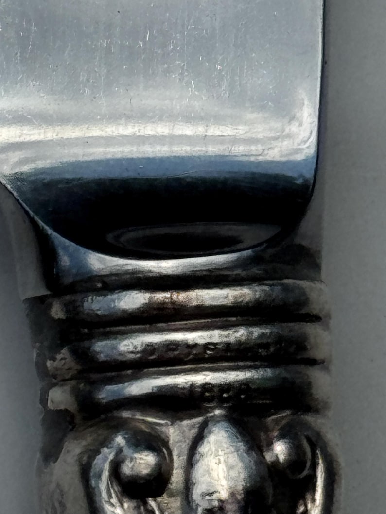 May include: Close-up of a silver knife with a reflective blade. The handle has textured rings and a decorative base. The word "STERLING" is visible on the handle.