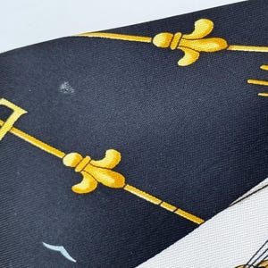 Puede incluir: Un pa&ntilde;uelo de seda azul marino con un dise&ntilde;o n&aacute;utico. El pa&ntilde;uelo presenta dise&ntilde;os de flor de lis y l&iacute;neas doradas sobre un fondo oscuro. Se ve una secci&oacute;n blanca con un dise&ntilde;o de barco.