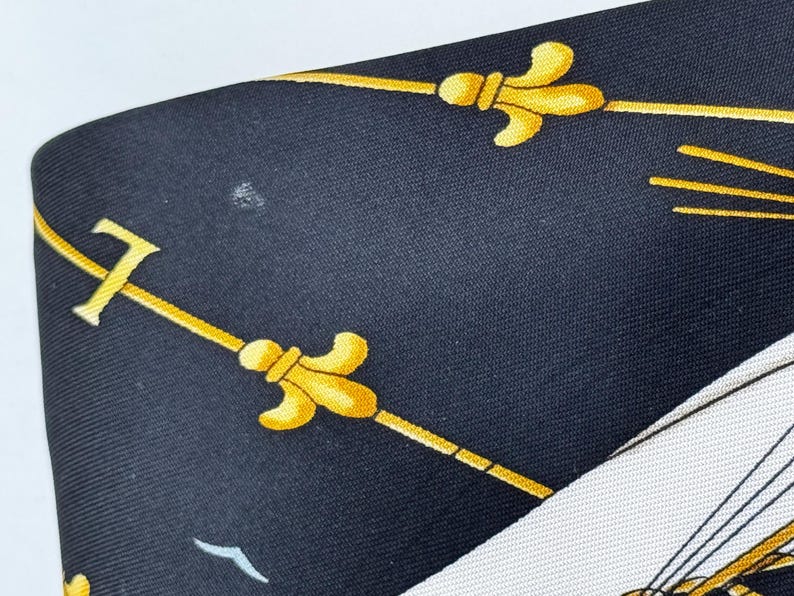 Puede incluir: Un pa&ntilde;uelo de seda azul marino con un dise&ntilde;o n&aacute;utico. El pa&ntilde;uelo presenta dise&ntilde;os de flor de lis y l&iacute;neas doradas sobre un fondo oscuro. Se ve una secci&oacute;n blanca con un dise&ntilde;o de barco.