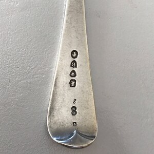 Może przedstawiać: Zbliżenie srebrnej łyżki z zaokrągloną miseczką i długim, wąskim trzonkiem. Trzonek posiada kilka stempli, w tym literę "B" i cyfrę "8". Łyżka ma wypolerowaną, odbijającą światło powierzchnię.