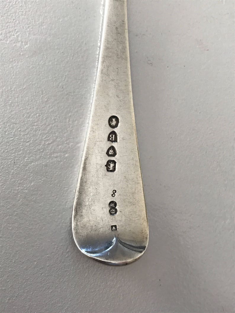 Może przedstawiać: Zbliżenie srebrnej łyżki z zaokrągloną miseczką i długim, wąskim trzonkiem. Trzonek posiada kilka stempli, w tym literę "B" i cyfrę "8". Łyżka ma wypolerowaną, odbijającą światło powierzchnię.