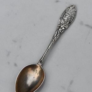 May include: A silver-toned metal spoon featuring an embossed handle design of a person wearing a feathered headdress. The handle also displays intricate botanical details, including corn stalks. The bowl of the spoon has a darkened, tarnished finish.