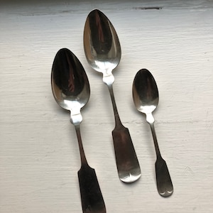 May include: Three silver spoons of varying sizes, with rounded bowls and ornate handles. The spoons are arranged in a row, with the largest spoon at the top and the smallest spoon at the bottom.