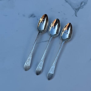 May include: Three antique silver spoons with oval bowls and ornate, engraved handles. The spoons are arranged diagonally, reflecting light. Each handle features an engraved design and initials. The spoons are likely for serving or dining.