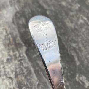 May include: Close-up of the top of a silver spoon with an engraved design of two crowns and a stylized arrow.