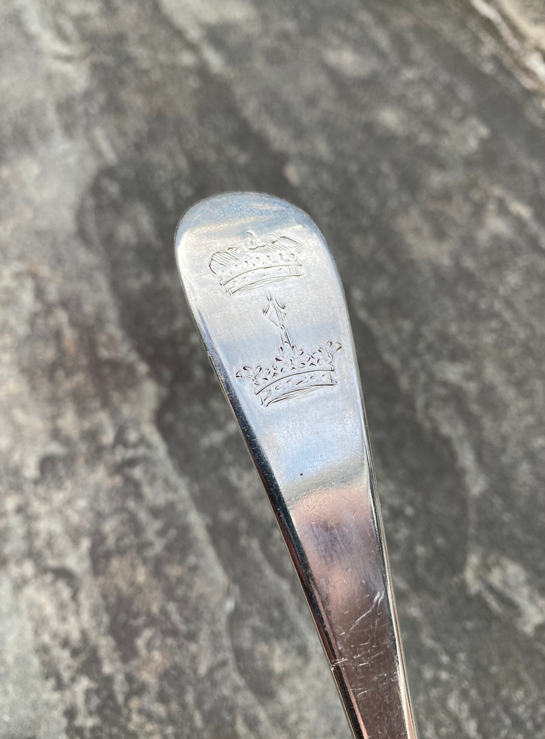 May include: Close-up of the top of a silver spoon with an engraved design of two crowns and a stylized arrow.