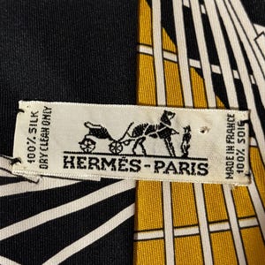 Puede incluir: Pa&ntilde;uelo de seda con un dise&ntilde;o geom&eacute;trico en negro, blanco y dorado. El pa&ntilde;uelo presenta el logotipo "HERM&Egrave;S-PARIS" y texto que indica que es 100% seda y est&aacute; hecho en Francia.