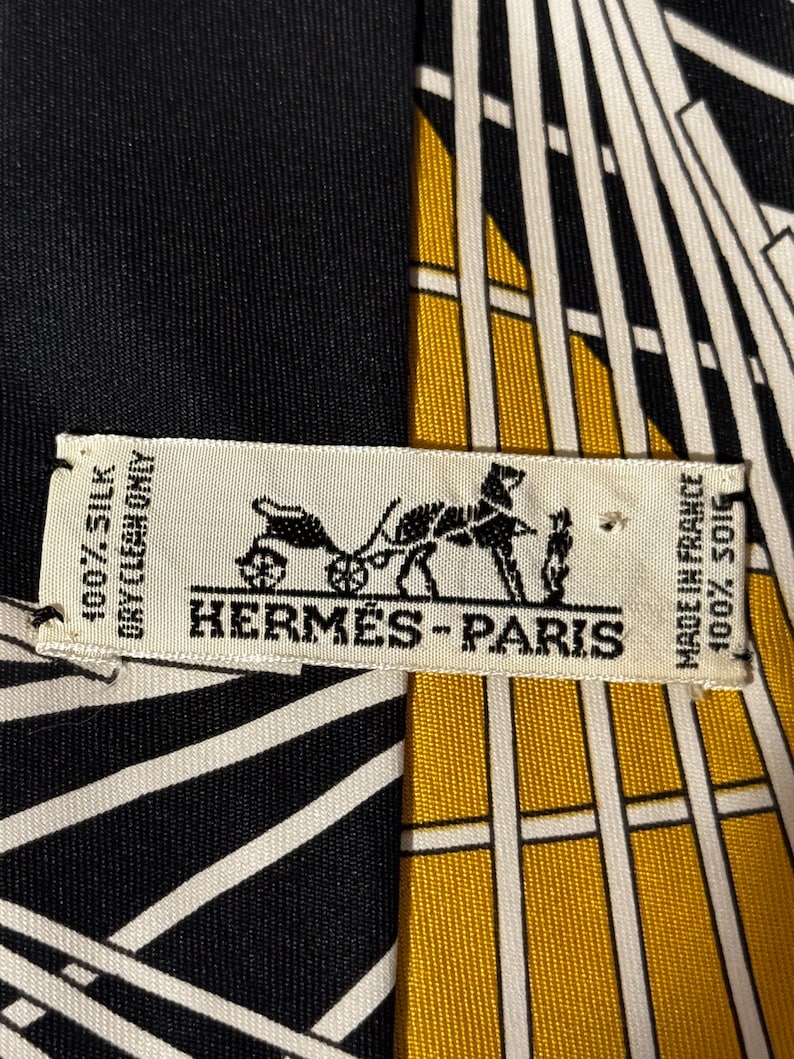 Puede incluir: Pa&ntilde;uelo de seda con un dise&ntilde;o geom&eacute;trico en negro, blanco y dorado. El pa&ntilde;uelo presenta el logotipo "HERM&Egrave;S-PARIS" y texto que indica que es 100% seda y est&aacute; hecho en Francia.