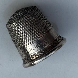 Könnte beinhalten: Ein silberner Fingerhut mit einem Wabenmuster auf der Oberseite und dem Wort "STERLING" auf dem Band eingraviert. Der Fingerhut hat ein dekoratives Blumendesign um die Basis.