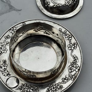 Op de afbeelding: Een zilveren botervloot met deksel, versierd met een bloemmotief. De schaal is rond met een centraal compartiment en een decoratieve rand. Het deksel heeft een kleine knop en past bij het bloemmotief. De schaal staat op een grijze ondergrond.