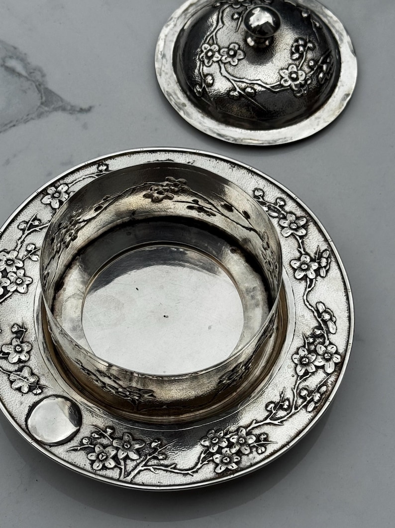 Op de afbeelding: Een zilveren botervloot met deksel, versierd met een bloemmotief. De schaal is rond met een centraal compartiment en een decoratieve rand. Het deksel heeft een kleine knop en past bij het bloemmotief. De schaal staat op een grijze ondergrond.