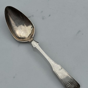 Può includere: Un cucchiaio d'argento con una coppa ovale e un manico lungo e sottile. Il manico presenta un motivo decorativo alla base della coppa e un motivo a conchiglia all'estremità. Il cucchiaio è probabilmente vintage o antico.
