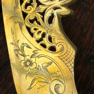 May include: A gold-toned metal butter knife with an intricate floral design. The knife has a curved handle and a pointed blade.
