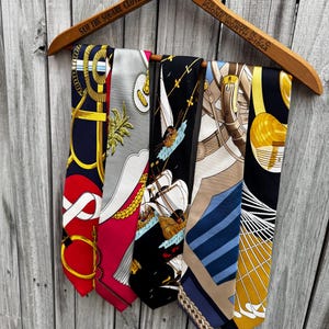 Puede incluir: Un colgador de madera exhibe cinco corbatas de seda con diversos dise&ntilde;os. Las corbatas presentan dise&ntilde;os en rojo, azul marino, negro y dorado, con motivos n&aacute;uticos y geom&eacute;tricos. El colgador tiene el texto "SAM THE SQUARE CLOTHIER" y "128 PARK ROW, N.Y.C."