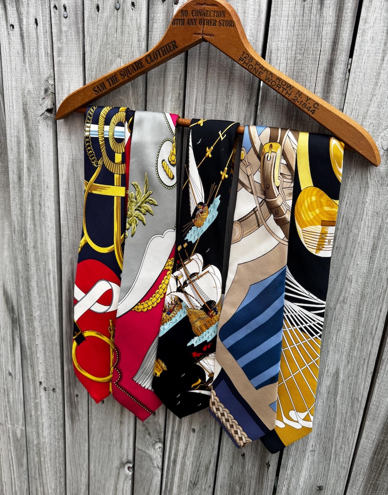 Puede incluir: Un colgador de madera exhibe cinco corbatas de seda con diversos dise&ntilde;os. Las corbatas presentan dise&ntilde;os en rojo, azul marino, negro y dorado, con motivos n&aacute;uticos y geom&eacute;tricos. El colgador tiene el texto "SAM THE SQUARE CLOTHIER" y "128 PARK ROW, N.Y.C."