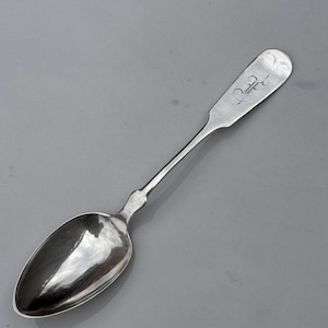 May include: A silver spoon with a long handle and an oval bowl. The handle has a decorative curve near the bowl and a rounded end with an engraved design. The spoon is set against a grey background.