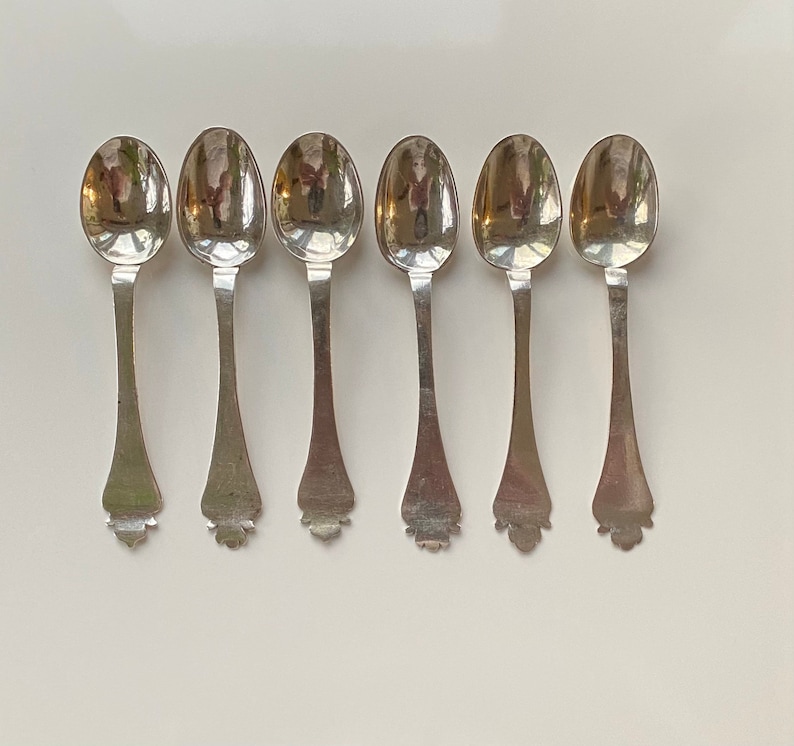 May include: Six silver teaspoons with ornate handles. The spoons are arranged in a row, with the handles facing the viewer.