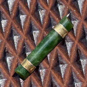 以下が含まれることがあります： 両端に金色の金属バンドが付いた円筒形のダークグリーンの翡翠ペンダント。金属バンドには葉の模様が刻印されています。ペンダントは滑らかで光沢のある仕上げで、真っ直ぐな細長いチューブ状の形状をしています。