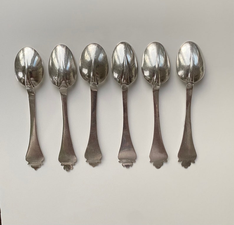 May include: Six silver teaspoons with ornate handles. The spoons are arranged in a row, with the handles facing the viewer.