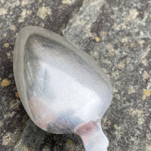 May include: A silver spoon with a rounded bowl and a simple, ornate handle. The spoon is lying on a textured, gray surface.