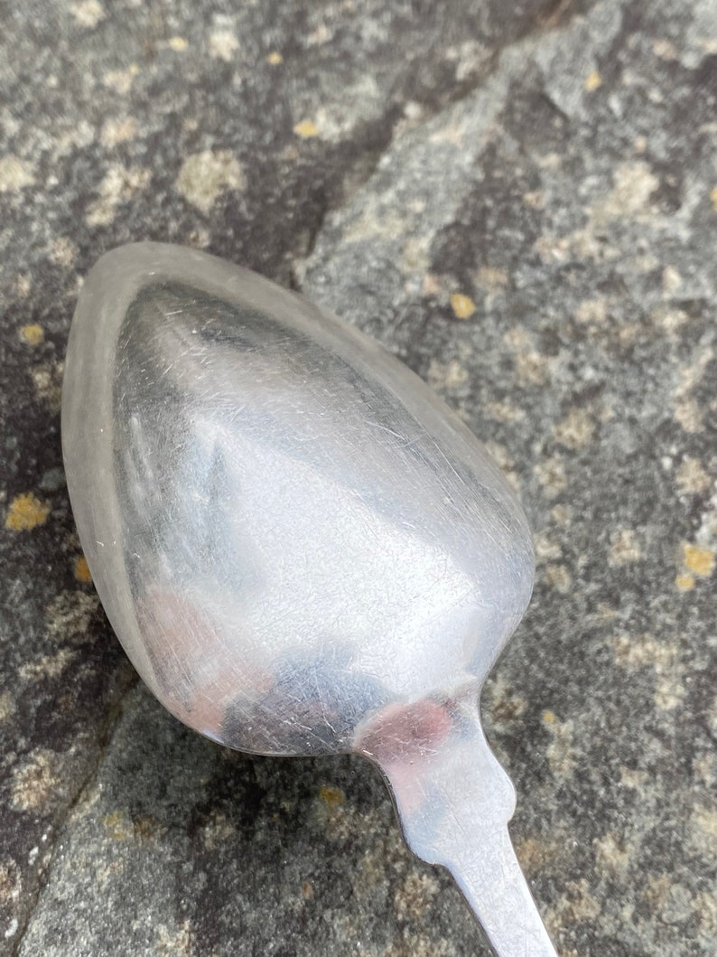 May include: A silver spoon with a rounded bowl and a simple, ornate handle. The spoon is lying on a textured, gray surface.