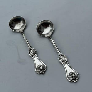 May include: Two antique silver spoons with round bowls and ornate handles. The handles feature a floral design with a leaf motif. The spoons are displayed on a gray background.