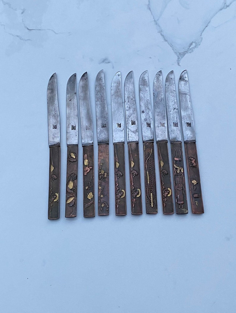 K&ouml;nnte beinhalten: Set aus 10 silbernen Messern mit verzierten Griffen aus Kupfer und Gold. Jeder Griff hat ein anderes Design mit floralen und geometrischen Mustern.