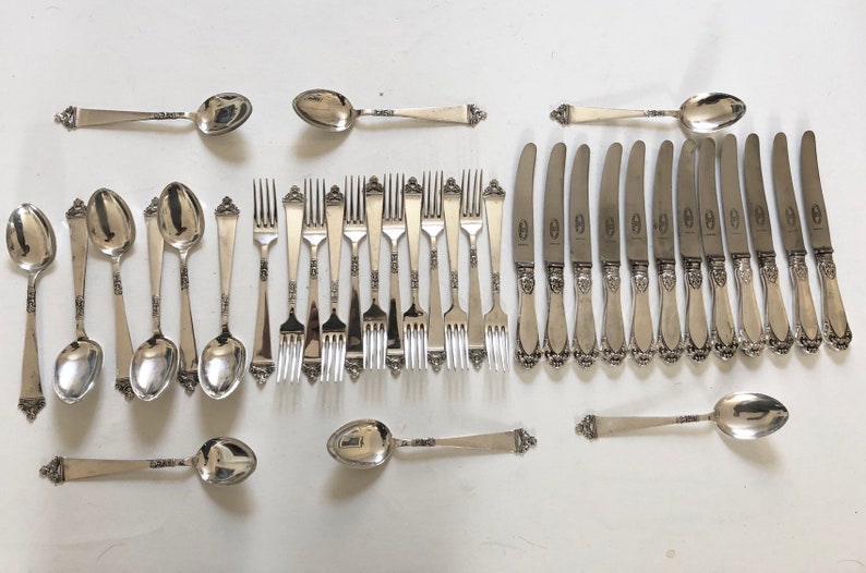 Puede incluir: Un juego de cubiertos de plata, que incluye cucharas, tenedores y cuchillos. Los cubiertos tienen un dise&ntilde;o adornado y est&aacute;n dispuestos en filas sobre una superficie blanca.