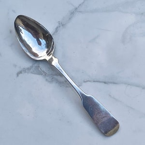 Puede incluir: Una cucharita de plata con un cuenco redondeado y un mango simple y elegante. El mango tiene un pequeño diseño grabado.