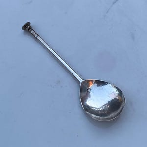 Può includere: Un cucchiaio d'argento con un manico lungo e sottile e una ciotola arrotondata. Il manico ha un pomo decorativo in cima.