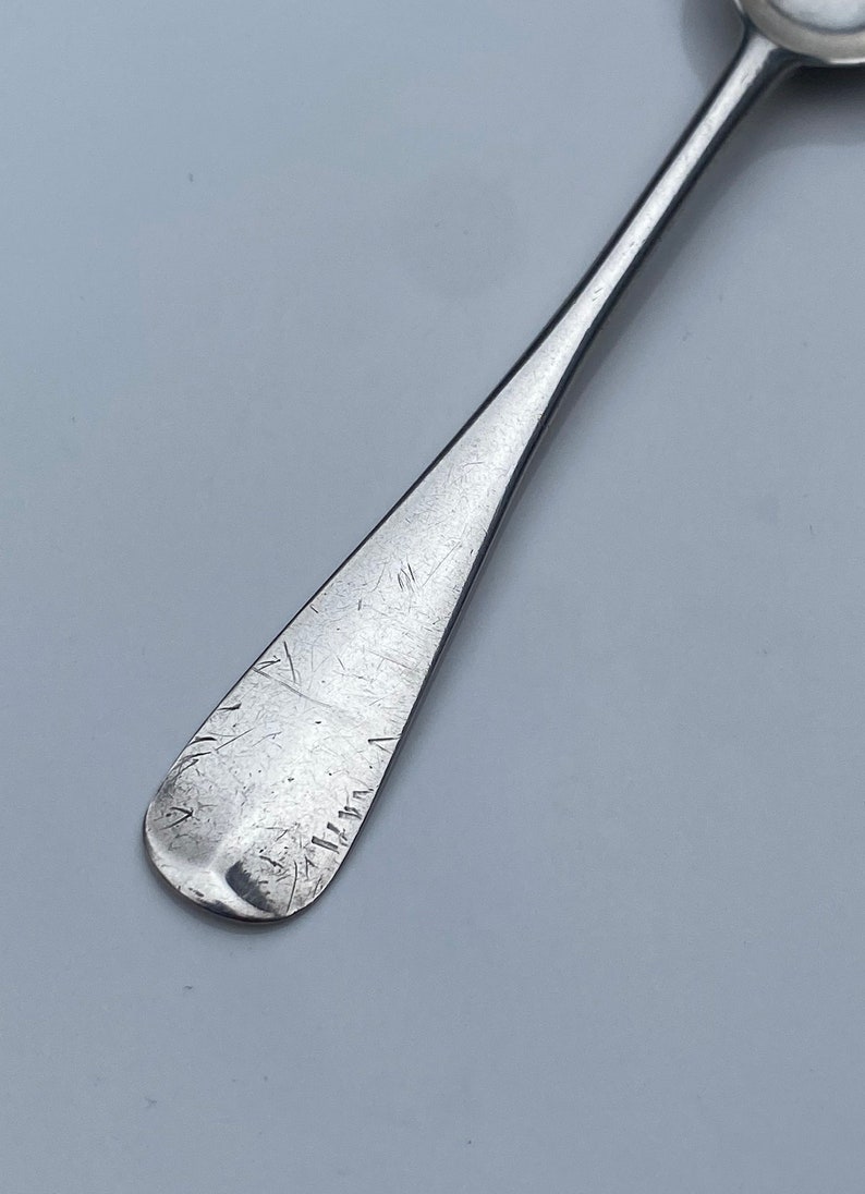 K&ouml;nnte beinhalten: Ein silberner Teel&ouml;ffel mit einer abgerundeten Schale und einem langen, schlanken Stiel. Der Stiel ist mit einer Reihe von Kratzern und Linien versehen.