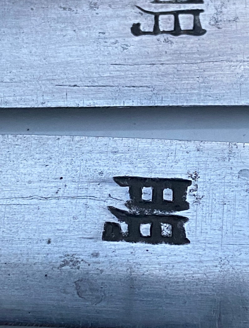 K&ouml;nnte beinhalten: Nahaufnahme einer silbernen Metalloberfl&auml;che mit einem schwarzen Kanji-Zeichen, das darauf eingepr&auml;gt ist. Das Zeichen ist eine stilisierte Darstellung eines dreist&ouml;ckigen Geb&auml;udes.