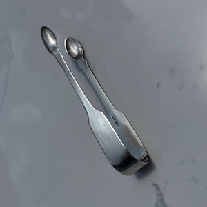 Può includere: Pinze per zucchero color argento con estremità a forma di cucchiaio. Le pinze hanno un design curvo e una cerniera al centro. Le pinze sono in metallo lucido, probabilmente vintage. Misurano circa 12 cm di lunghezza.