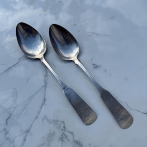 May include: Two antique silver spoons with oval bowls and long, slender handles. The handles have a decorative design near the bowl and an engraved initial. The spoons are displayed on a white marble surface.