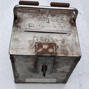 May include: A rectangular metal ballot box with a silver finish, featuring a top slot, a hinged lid with two brown hinges, and a metal handle. The surface has etched text reading LOCAL #521, BALLOTS, and HERE, along with a central metal latch.