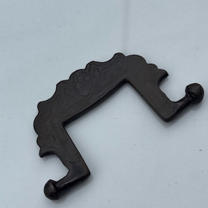 Könnte beinhalten: Ein dunkelbraunes, verziertes Metallobjekt mit einer gebogenen, eckigen Form. Der obere Rand weist ein dekoratives, gewelltes Design mit geätzten Details auf. An den Enden der abgewinkelten Arme befinden sich zwei abgerundete Knöpfe. Das Objekt scheint ein dekoratives Hardware-Teil zu sein.