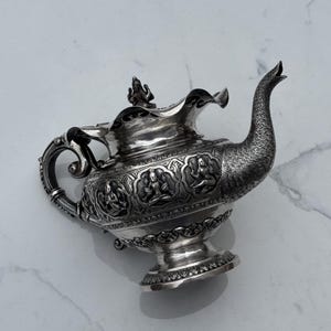 Könnte beinhalten: Eine silberne Teekanne mit aufwendigen floralen und figürlichen Verzierungen. Die Teekanne hat einen geriffelten Ausguss und einen Deckel mit einem Knauf.