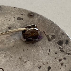 May include: Close-up of the back of a silver circular brooch with a pin and catch. The brooch has a textured surface and the word "OKO" is visible on the bottom.