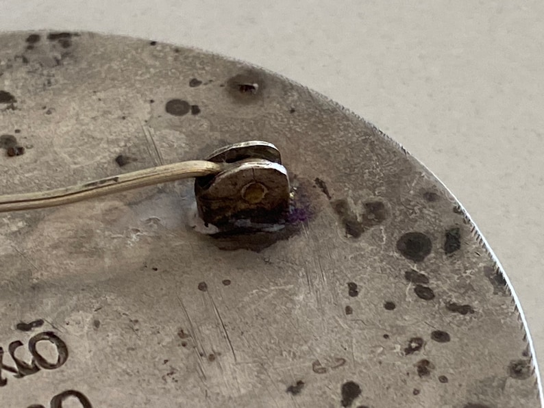May include: Close-up of the back of a silver circular brooch with a pin and catch. The brooch has a textured surface and the word "OKO" is visible on the bottom.