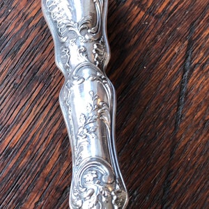 May include: A silver handle with intricate floral designs and a brass base. The handle is ornate and detailed, with a polished finish.
