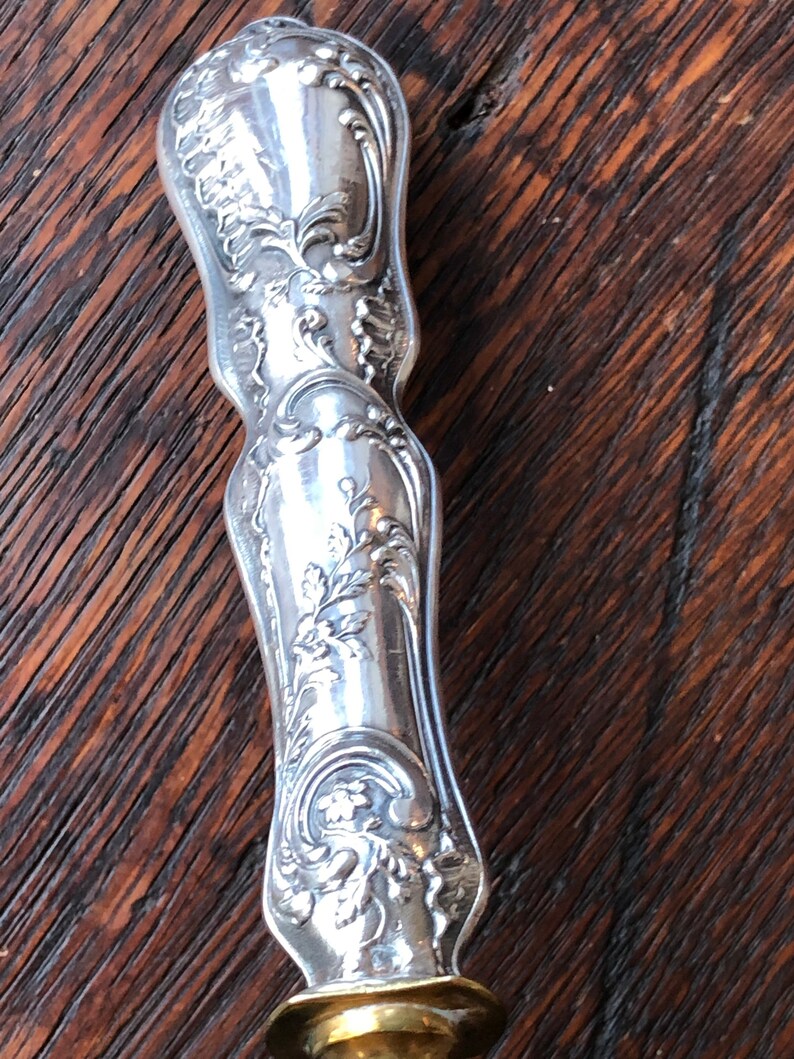 May include: A silver handle with intricate floral designs and a brass base. The handle is ornate and detailed, with a polished finish.