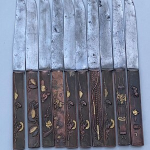 K&ouml;nnte beinhalten: Set mit 10 japanischen Messern mit verzierten Griffen. Die Griffe sind aus Metall und weisen komplizierte Designs in Gold und Kupfer auf. Die Klingen sind aus Stahl und haben eine polierte Oberfl&auml;che.