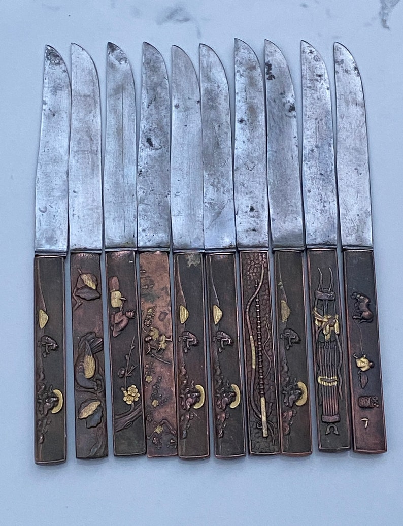 K&ouml;nnte beinhalten: Set mit 10 japanischen Messern mit verzierten Griffen. Die Griffe sind aus Metall und weisen komplizierte Designs in Gold und Kupfer auf. Die Klingen sind aus Stahl und haben eine polierte Oberfl&auml;che.