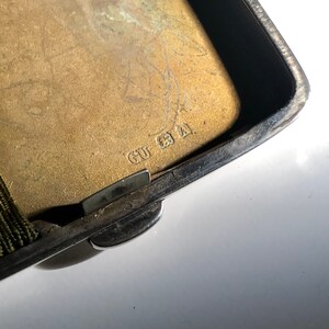Pu&ograve; includere: Primo piano di un porta sigarette in argento con un coperchio incernierato. Il porta sigarette ha un elastico verde e l'iscrizione "GU 21" sul fondo.