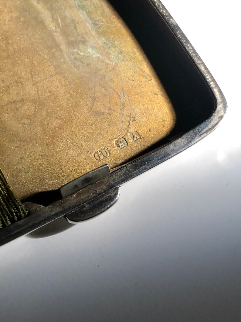 Pu&ograve; includere: Primo piano di un porta sigarette in argento con un coperchio incernierato. Il porta sigarette ha un elastico verde e l'iscrizione "GU 21" sul fondo.