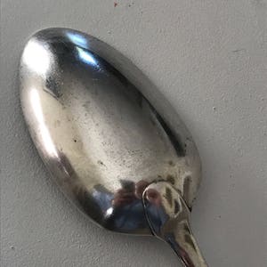 Może przedstawiać: Zbliżenie srebrnej łyżki z zaokrągloną miseczką i długim, smukłym trzonkiem. Łyżka odbija otoczenie, ukazując błyszczącą, wypolerowaną powierzchnię. Łyżka jest prawdopodobnie używana do jedzenia lub serwowania potraw.